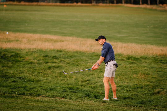 Male golfer follows through on a swing from the rough on a lush golf course. Wearing casual sporty attire and focused on accuracy during the game. - Powered by Adobe