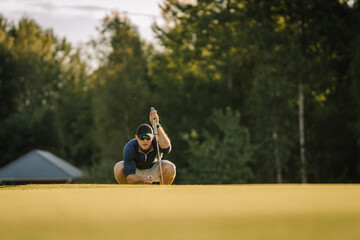 Male golfer squats down on the green, lining up his putt with intense focus. Club in one hand, he assesses the shot on a sunny course with forest backdrop.