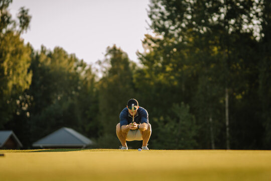 Male golfer squats low on the green, aligning his putter and aiming at the ball with focus. Surrounded by trees and soft evening light on a quiet course.