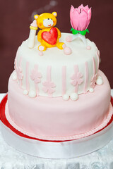 A two-tier pink cake decorated with fondant flowers, a teddy bear holding a heart, and a pink lotus candle topper for a children’s birthday celebration.
