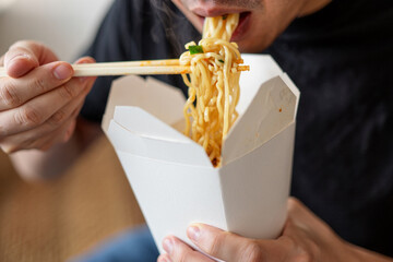 Man eating curly noodles with chopsticks from white takeout container, with blurred background emphasizing food and action.