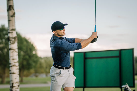 Male golfer finishes his swing at a practice range. Wearing sporty attire and sunglasses, he stands confidently with trees and a target board in the background.