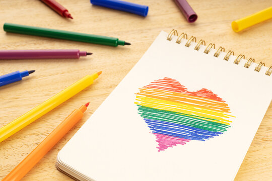 Notebook with LGBT rainbow flag heart shaped drawing and marker pens on wooden table - Powered by Adobe