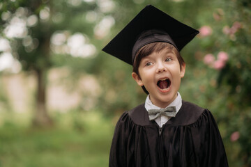 Portrait of emotional boy-graduate in graduation dress and hat on green background, spring. Children's education concept