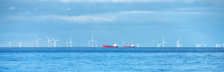 A vast expanse of ocean stretches under a bright blue sky, dotted with white wind turbines gently rotating in the distance. Two ships travel across the calm sea, adding a touch of vibrancy.
