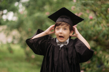 Portrait of emotional boy-graduate in graduation dress and hat on green background, spring. Children's education concept