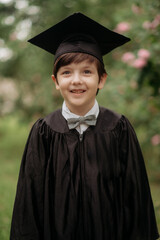 Portrait of emotional boy-graduate in graduation dress and hat on green background, spring. Children's education concept