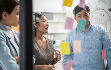 Asian business team in a creative meeting, collaborating and sharing ideas using colorful notes on a glass wall. A modern teamwork setting showing brainstorming, innovation, and planning in office