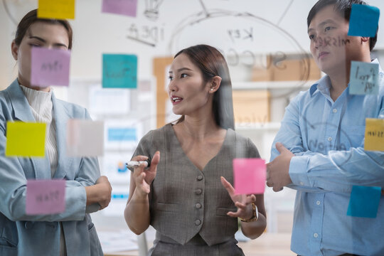 Asian business team in a creative meeting, collaborating and sharing ideas using colorful notes on a glass wall. A modern teamwork setting showing brainstorming, innovation, and planning in office
