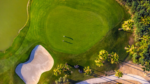 Golfers playing on lush green course with sand trap and palm trees, aerial view