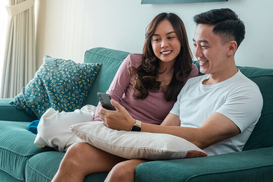 Young Asian couple sitting on green sofa at home, smiling while looking at smartphone together. Woman in pink top, man in white t-shirt with smartwatch, cozy indoor living room setting