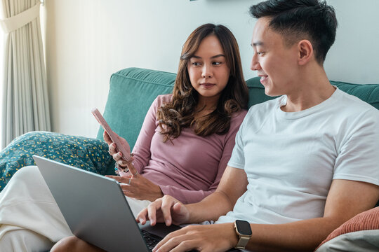 Young Asian couple sitting on green sofa at home, smiling while using laptop and smartphone together. Woman wears pink top, man in white t-shirt with smartwatch, cozy indoor background - Powered by Adobe