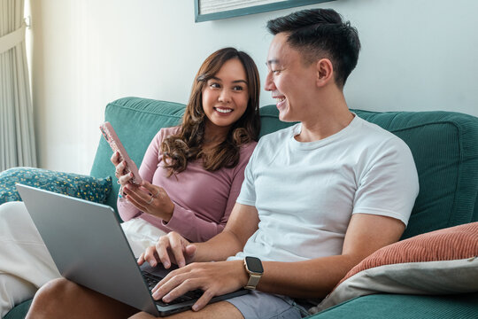 Young Asian couple sitting on green sofa at home, smiling while using laptop and smartphone together. Woman wears pink top, man in white t-shirt with smartwatch, cozy indoor background