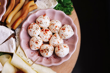 Mozzarella cheese balls with paprika spice in pink bowl. Wooden board with assorted cheeses and smoked sticks. Fresh lettuce and rustic snack background. Tasty dairy appetizer. Gourmet food serving.