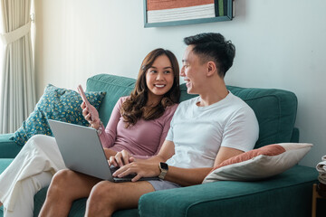 Young Asian couple sitting on green sofa at home, smiling while using laptop and smartphone together. Woman wears pink top, man in white t-shirt with smartwatch, cozy indoor background