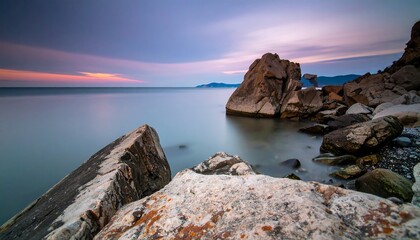 Calm coastal rocks at sunrise, bathed in serene pastels, showcase tranquil water and dramatic rock formations.