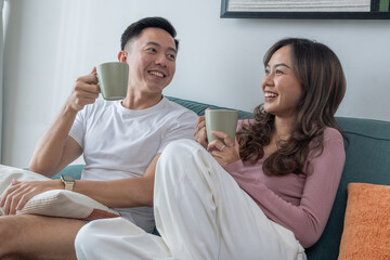 Smiling young Asian couple sitting on green sofa at home, enjoying hot drinks together. Woman in pink top, man in white t-shirt with smartwatch, cozy indoor living room background