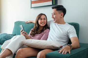 Obraz premium Smiling young Asian couple sitting on green sofa at home, looking at each other while holding smartphone. Woman in pink top, man in white t-shirt with smartwatch, cozy indoor setting