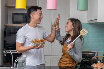 Asian couple smiling in kitchen, giving a high five while holding fried chicken and spatula, celebrating teamwork and happiness in cooking at home together