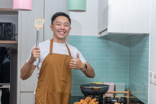Asian man in apron smiling in the kitchen, holding a spatula and making an OK hand gesture, standing near stove with frying pan, fried chicken, and eggs on the counter