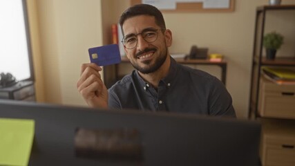 Young man holding credit card smiling at computer in office suggesting online shopping positive financial transaction success indoors workspace relaxed. - Powered by Adobe