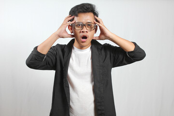 Portrait of a shocked young adult man with glasses, wearing a black shirt over a white t-shirt, clutching his head with both hands and an open mouth, shot in a studio against a plain white background