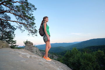 Naklejka premium Hiker woman standing on mountain footpath enjoying evening nature during her travel on wilderness trail. Lonely female traveler traversing high hilltop route. Healthy lifestyle concept