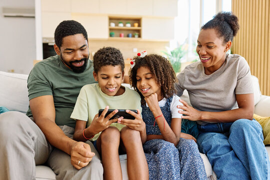 A family of four sits on a comfortable couch, engrossed in content on a mobile device. The parents and two kids share smiles and laughter in their bright living room. - Powered by Adobe