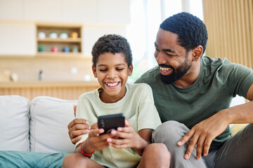 Teenage boy sitting at sofa with his father and using phone. Father and son bonding and having fun at home