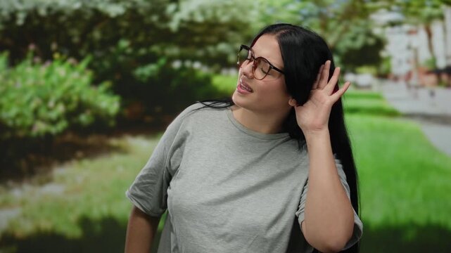 Woman standing in a park wearing glasses and a grey shirt with a hand gesture under the leafy green trees.