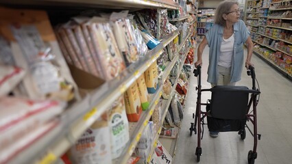 An elderly woman uses her walker to shop in a grocery store, carefully navigating the aisles while examining products on the shelves.