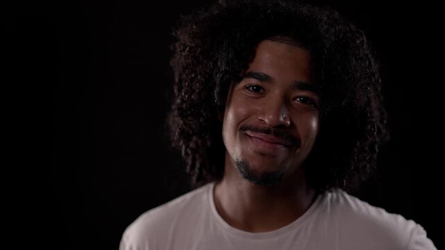 Portrait of a cheerful young latin African-American man with curly hair and goatee smiling on black background