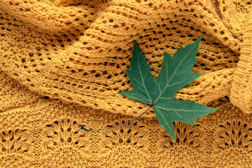 Green maple leaf lying on warm knitted yellow fabric creating cozy autumn seasonal background. Top view