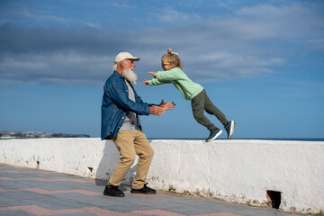Playful grandfather with white beard catching his cheerful blond grandson in green sweatshirt and sunglasses as the boy jumps fearlessly from a wall by the seaside promenade