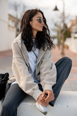 Stylish young woman with long dark hair, wearing sunglasses, oversized beige hoodie and casual outfit, sitting outdoors on a white wall in a modern residential area