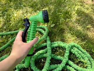 Hand holding green expandable garden hose with spray nozzle on grass in summer garden
