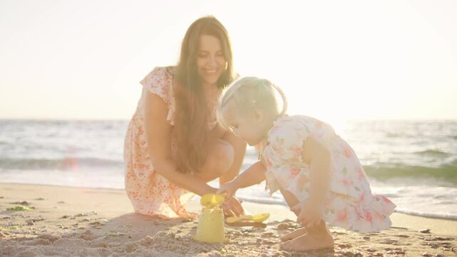 A joyful mother helps her young daughter build a sandcastle on a sunny beach, with ocean waves gently lapping at the shore during late afternoon light.