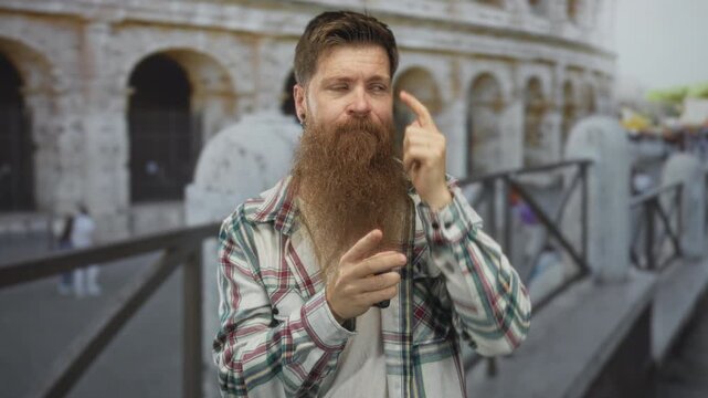 Man with long ginger beard points finger to temple while standing before sunlit ancient colosseum ruins; quiet reflection.