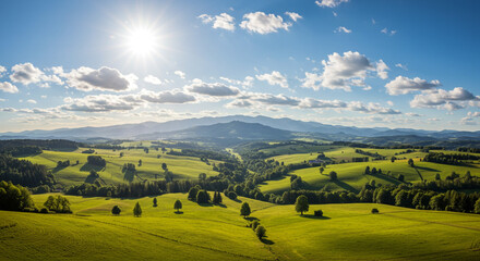 Obraz premium A scenic view of rolling green hills under a bright sun and a cloudy blue sky with distant mountains
