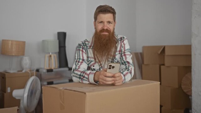 Man holds smartphone in both hands atop a cardboard box in a building under transition with stacked moving cartons and unpacked decor; anticipation.