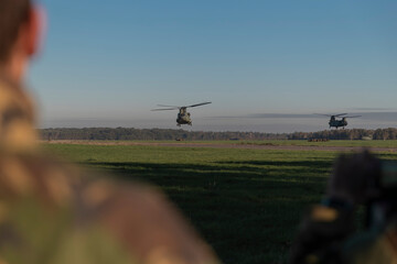 Multiple military Chinook helicopters flying over field with soldiers observing from ground during...