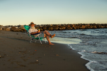 Elderly man with straw hat and white beard sitting comfortably in a beach chair near the ocean, holding orange juice, enjoying quiet sunset and peaceful retirement lifestyle in nature.