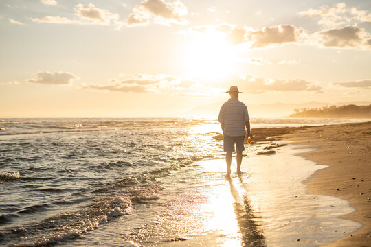 Elderly man in a straw hat walking barefoot along the beach shoreline at golden hour, holding a glass of orange juice and enjoying the peaceful sunset over the calm sea.