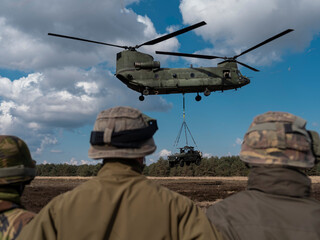 Military helicopter with soldiers observing sling load operations from ground positions with helmets