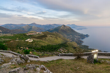Scenic view of the Maratea coast and surrounding mountains, taken from a high vantage point. The landscape features winding roads, lush greenery, rocky paths and the Tyrrhenian Sea
