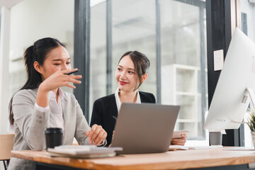 Two Asian businesswomen in formal suits discuss work at modern office desk with laptop, desktop computer, notebook, and coffee cup, showing teamwork and positive collaboration