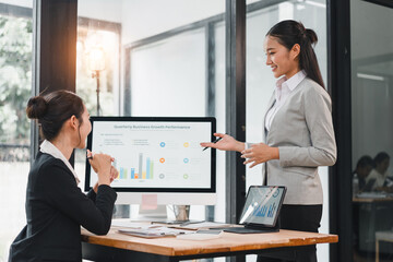 Two businesswomen discussing quarterly business growth performance with charts on computer and tablet in modern office, showing positive and engaged expressions