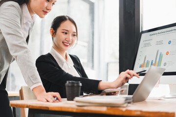 Two Asian businesswomen in formal suits discussing data analysis at modern office desk, smiling and collaborating on computer with business growth chart, teamwork and productivity
