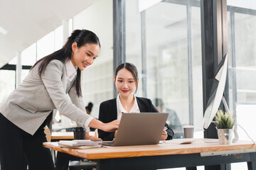 Two businesswomen Asian working together at modern office desk using laptop discussing project smiling professional teamwork bright workspace collaboration productivity positive atmosphere