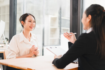 Two Asian women sitting at desk in modern office, one conducting job interview with clipboard, both smiling and engaged in positive conversation